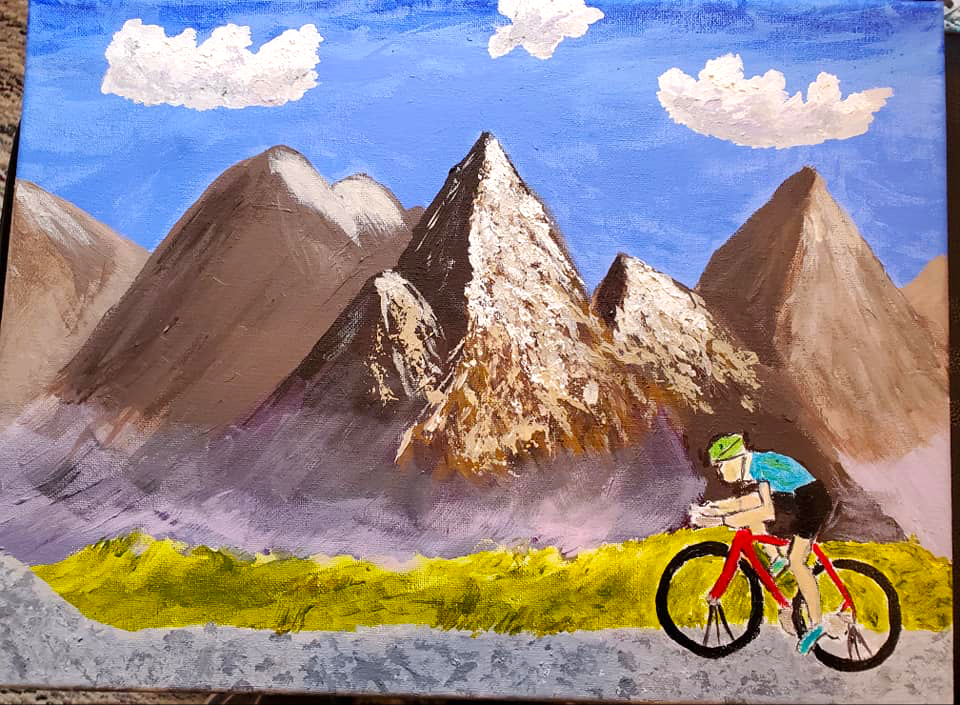 A bicyclist rides across a rugged landscape in front of ragged mountains with clouds high above in the blue sky