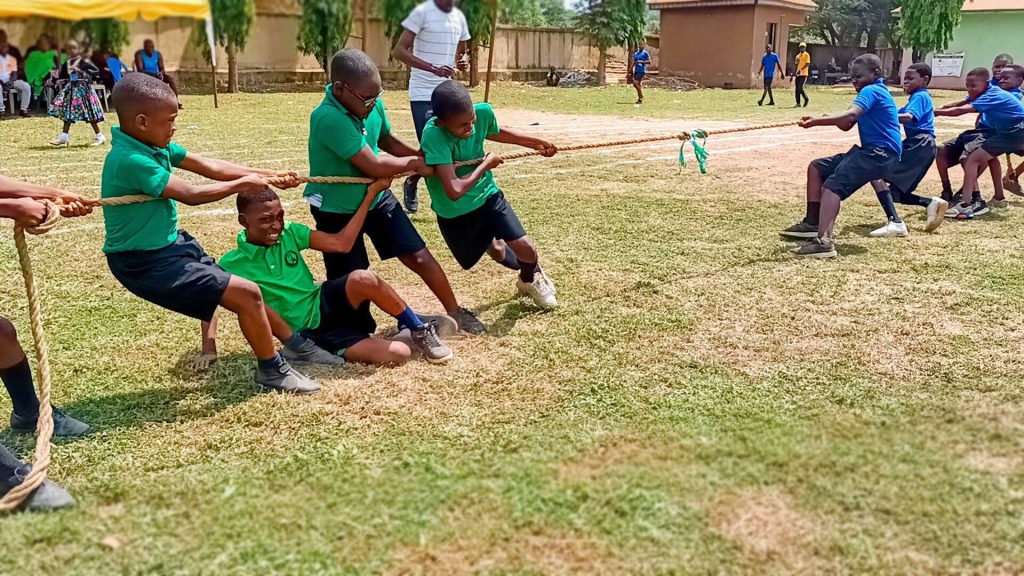 A photograph of Nigerian school children playing tug of war. The team on the left is wearing green shirts and black shorts and the team on the right is wearing blue shirts and black shorts. The rope they are pulling against has a green ribbon tied in the center. Each child is pulling forcefully against the rope and has a facial expression demonstrating exertion. The feet of each child are pushing against the grassy ground.