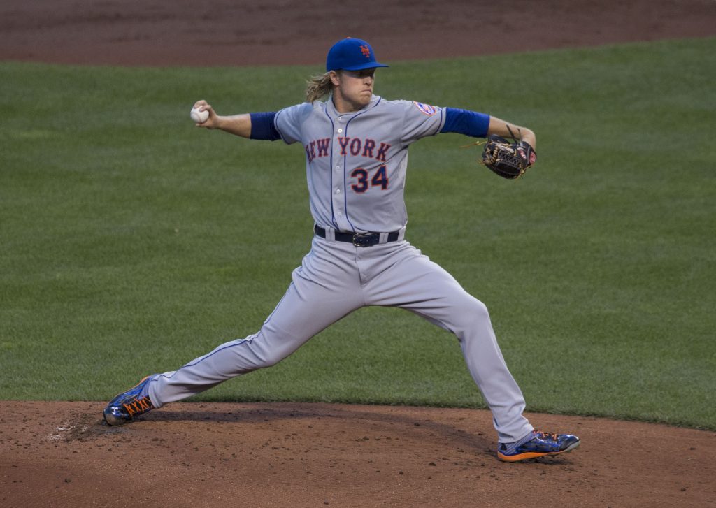 New York Mets pitcher Noah Syndergaard stands with arms outstretched, ready to pitch a baseball. His right hand holds the baseball as far back as possible. His left hand has a pitcher's mit. He is standing on the pitching mound in a baseball stadium.
