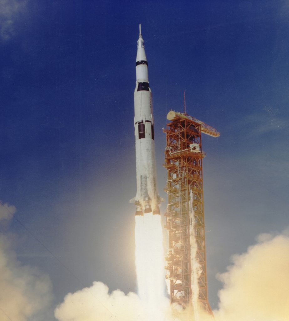 A 363 foot tall, 6,400,000 pound Saturn V rocket launches from Kennedy Space Center, Florida. It is propelled upwards into a clear blue sky by a jet of flames shooting out of the bottom boosters of the launch vehicle, creating clouds of smoke. The launch tower is a red steel framework.