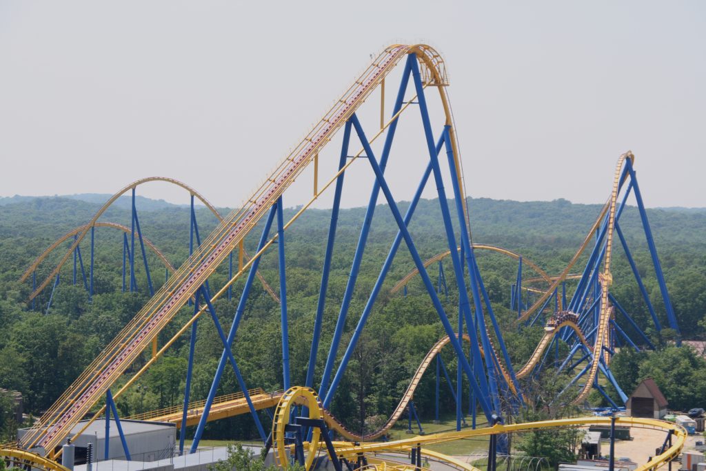 Photograph of a roller coaster with blue steel framework and a yellow track. The roller coaster rises up much taller than the surrounding trees, and in the background can be viewed a gray sky.