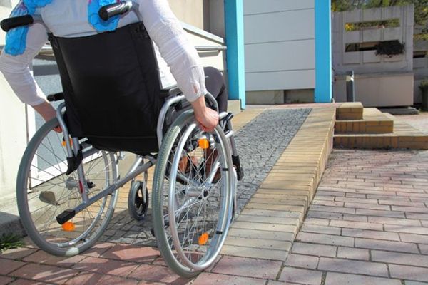 A wheelchair user is photographed from behind has they wheel up an outdoor brick ramp to access a building.