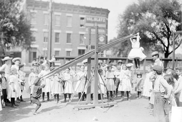 A black and white photograph of two children playing on a seesaw at the Webster School playground, with one of the children hanging by her hands from the raised end. Her feet dangle about a meter above the ground. A few dozen other schoolchildren and teachers look on in the background.