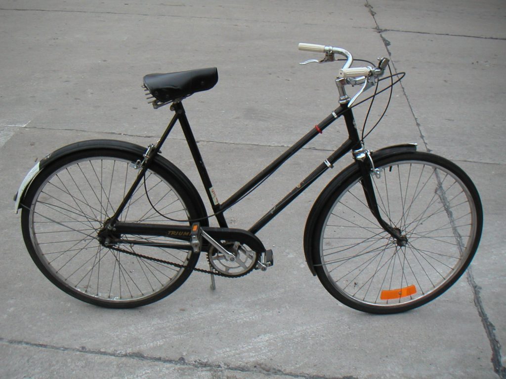 A black bicycle is propped upright by means of a kickstand on a concrete walkway.