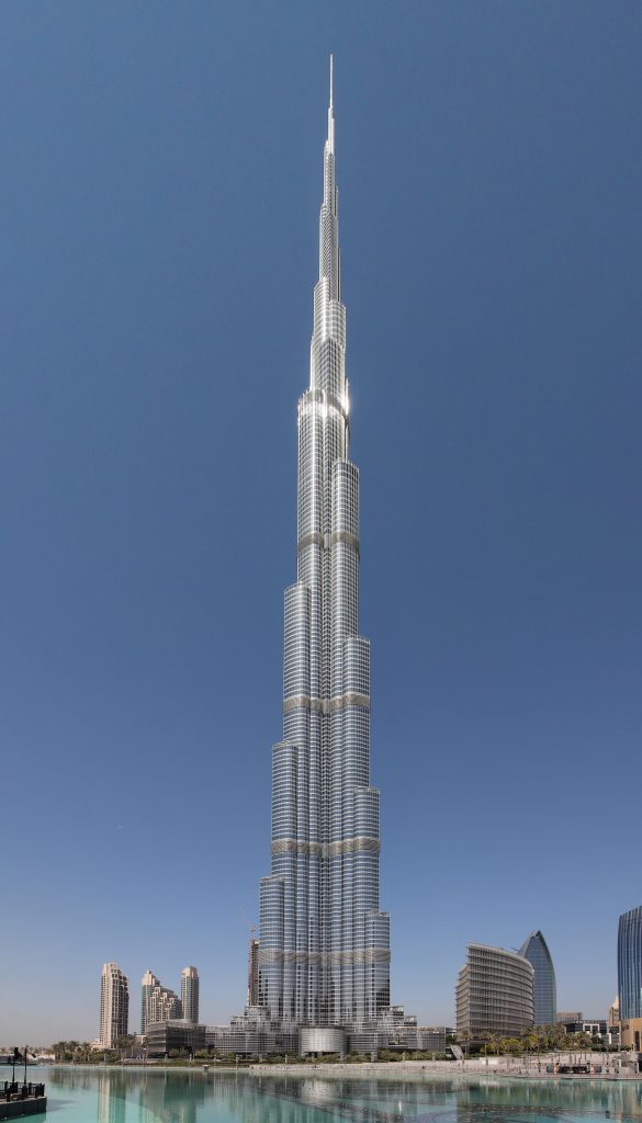 A photograph of the Burj Khalifa, a towering skyscraper. There are several other buildings nearby, much shorter than the Burj Khalifa. There is a cloudless blue sky in the background, and a waterway in the foreground of of the photograph.