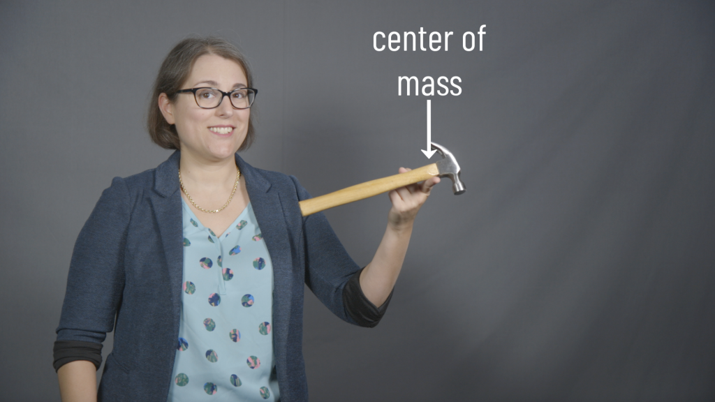 A photograph of Dr. Pasquale, a white lady with brown hair and glasses. She is balancing a hammer using one finger located near to the head of the hammer. An arrow over this point is labeled "center of mass." In the background is a gray curtain.