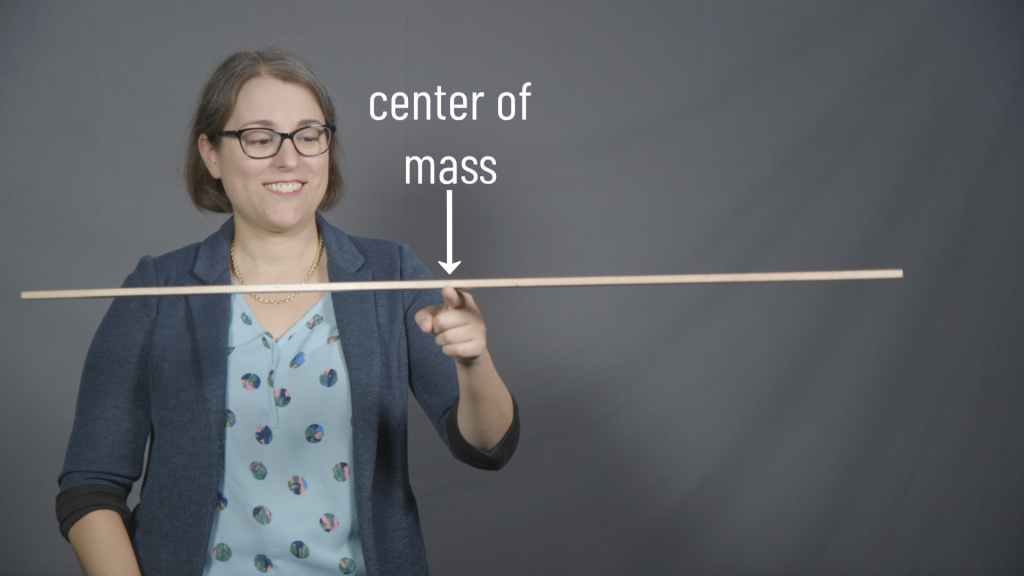 A photograph of Dr. Pasquale, a white lady with brown hair and glasses. She is balancing a meter stick using one finger located under the center of the meter stick. An arrow over this point is labeled "center of mass." In the background is a gray curtain.