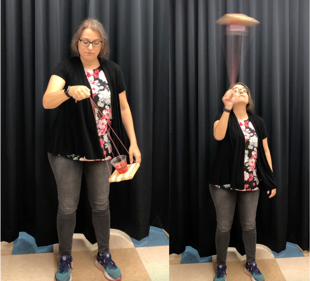 Two side-by-side photographs of Dr. Pasquale, a white lady with brown hair and glasses. On the left, she is holding a sling holding a cup as it is positioned at the bottom of a vertical circular path. On the right, she is holding the same sling while it is positioned at the top of a vertical circular path. There is water in the cup, dyed red. In both photographs, the water is situated in the bottom of the cup. In the background of both photographs is a black curtain.