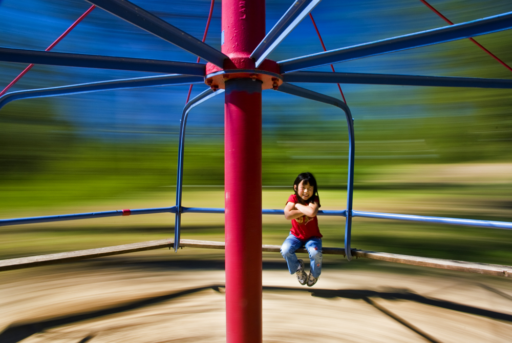 A small child sits on a playground merry-go-round. Because of the spinning of the merry-go-round, the background is blurry.