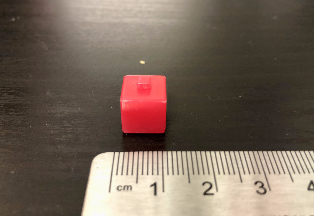 A photograph of a red plastic cube sitting on a black lab table. A metal ruler is positioned in the foreground for reference, displaying an edge length of 1 cm.