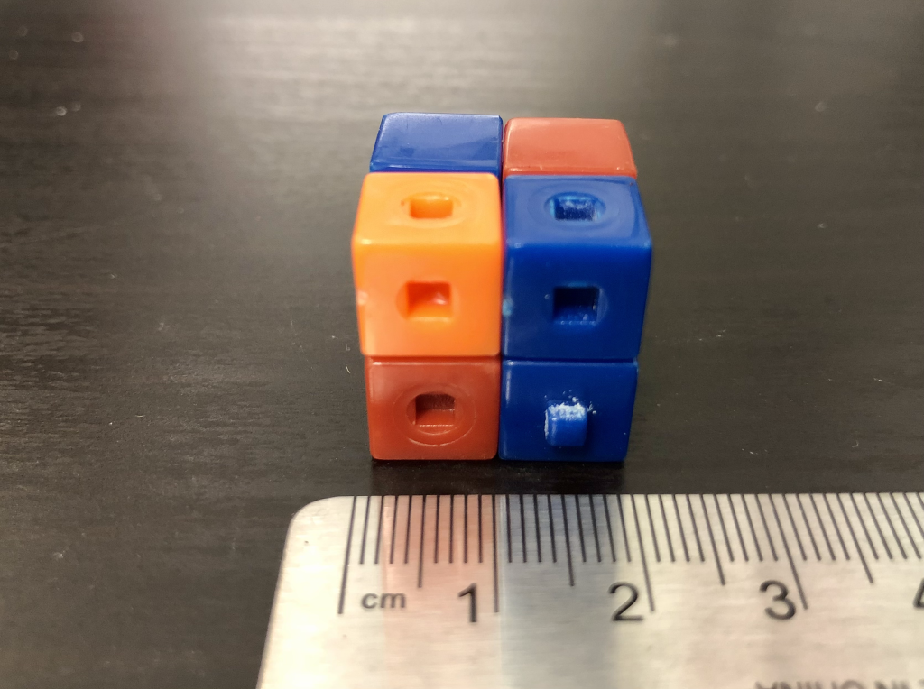 A photograph of eight different colored plastic cubes arranged together in a larger cube. The cube is sitting on a black lab table. A ruler is positioned in the foreground for reference, displaying an edge length of 2 cm.