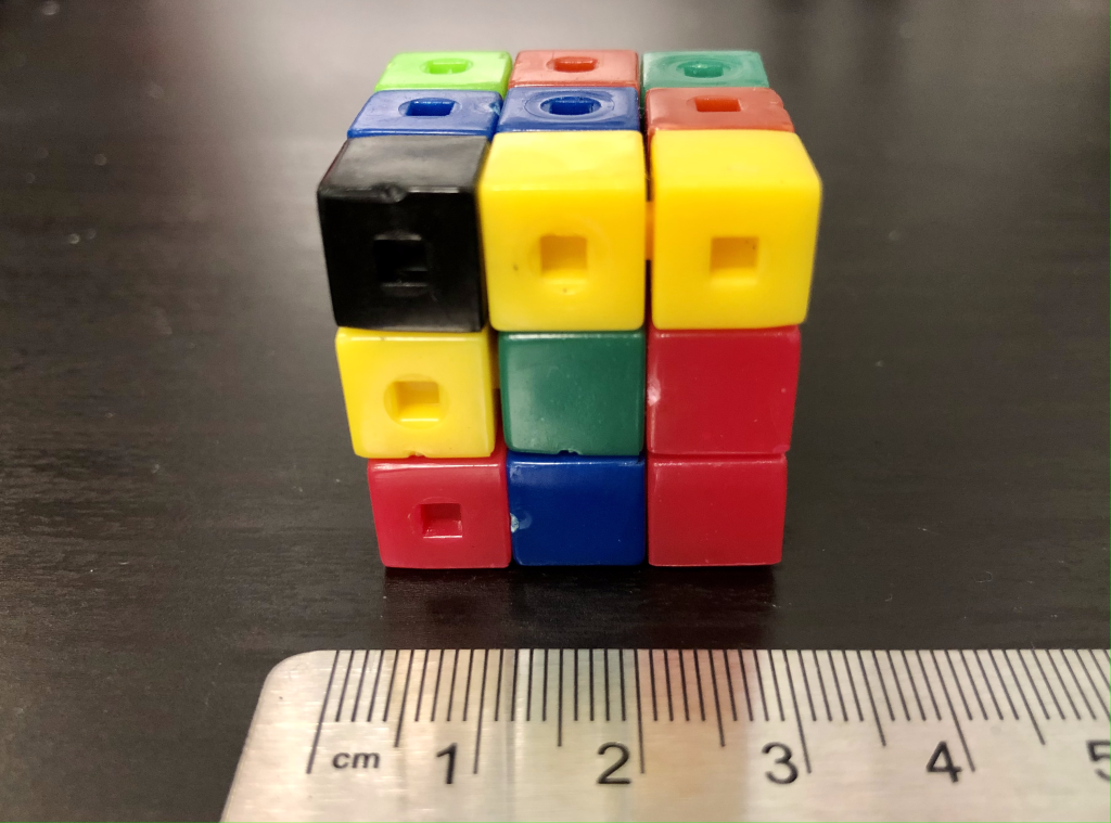 A photograph of 27 different colored plastic cubes arranged together in a larger cube. The cube is sitting on a black lab table. A ruler is positioned in the foreground for reference, displaying an edge length of 3 cm.