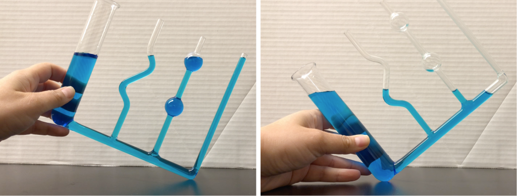 Two photographs of a glass container with many differently-shaped appendages. In both photographs, the container is filled with water dyed blue. Dr. Pasquale's hand is seen holding the container at different angles in each photograph. In both cases, the water level is completely horizontal.