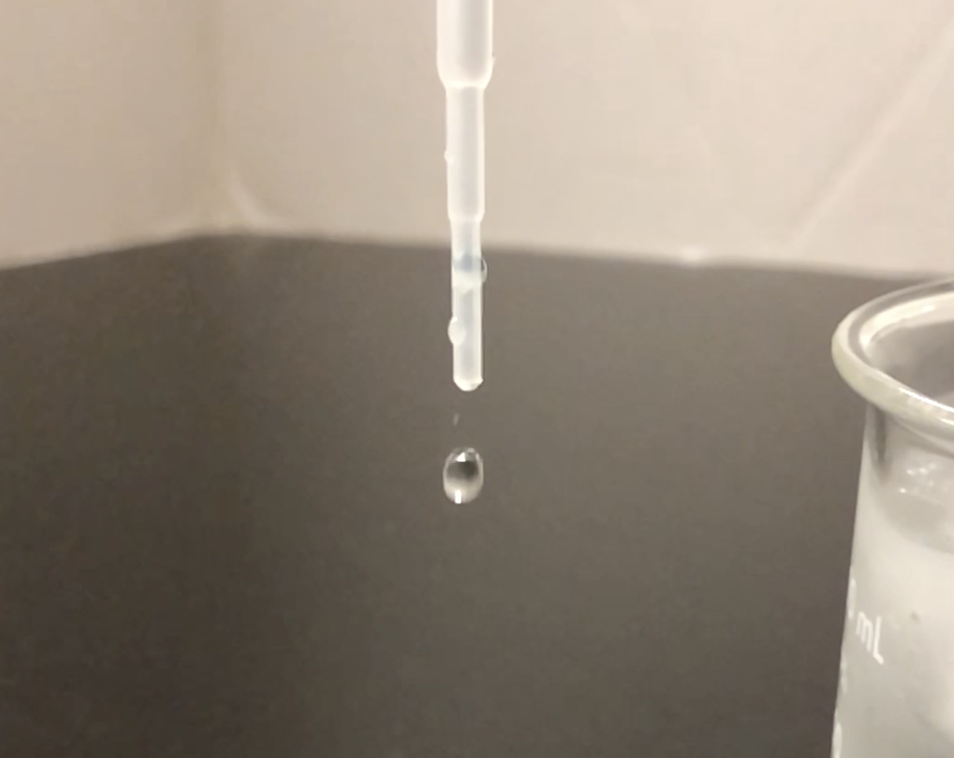 A photograph of a water droplet falling out of a plastic pipette. The water droplet is spherical in shape.