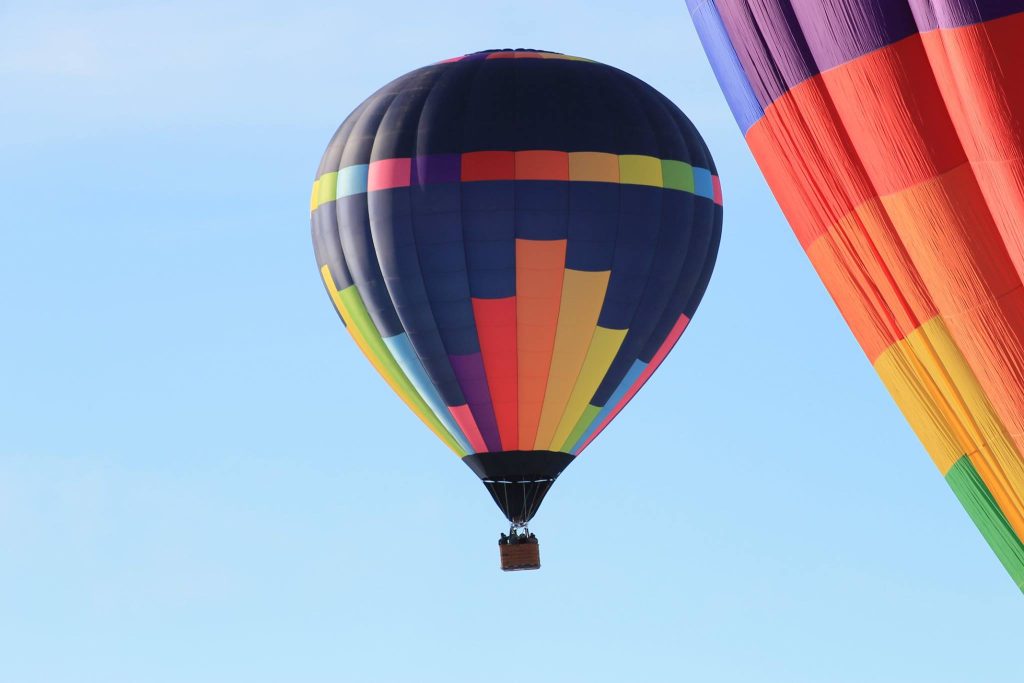 A hot air balloon with a rainbow colored envelope is seen lifting several people in the basket aloft.
