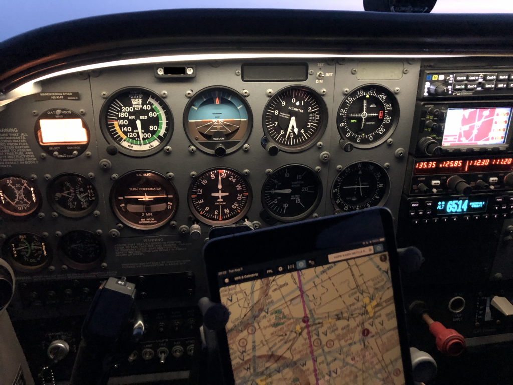 An airplane instrument panel consisting of various round gauges displaying altitude, airspeed, vertical speed, attitude, coordination, fuel level, heading, and fuel flow. In the foreground is an ipad displaying a route. To the right of the instrument panel are two radios and an aircraft transponder.