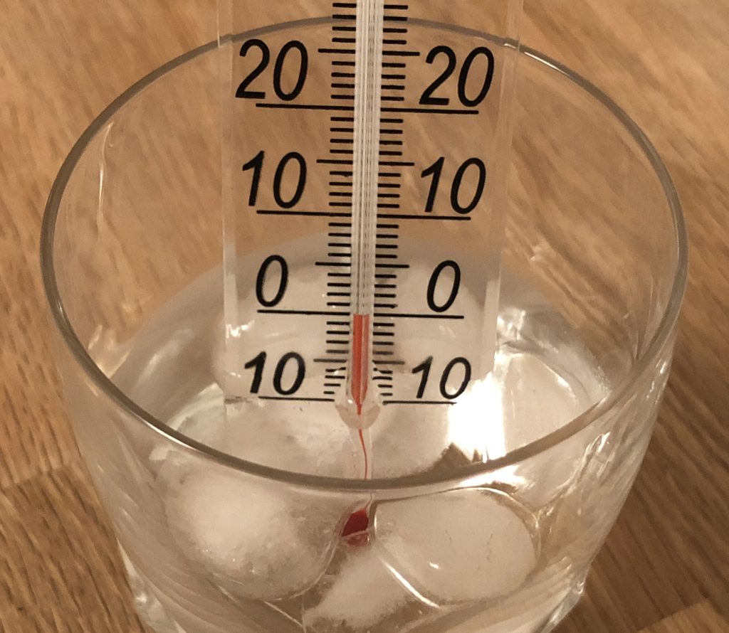 A photograph of a Celsius thermometer placed into a measuring cup filled with melting ice. The red dyed alcohol in the thermometer reads 0 degrees Celsius.