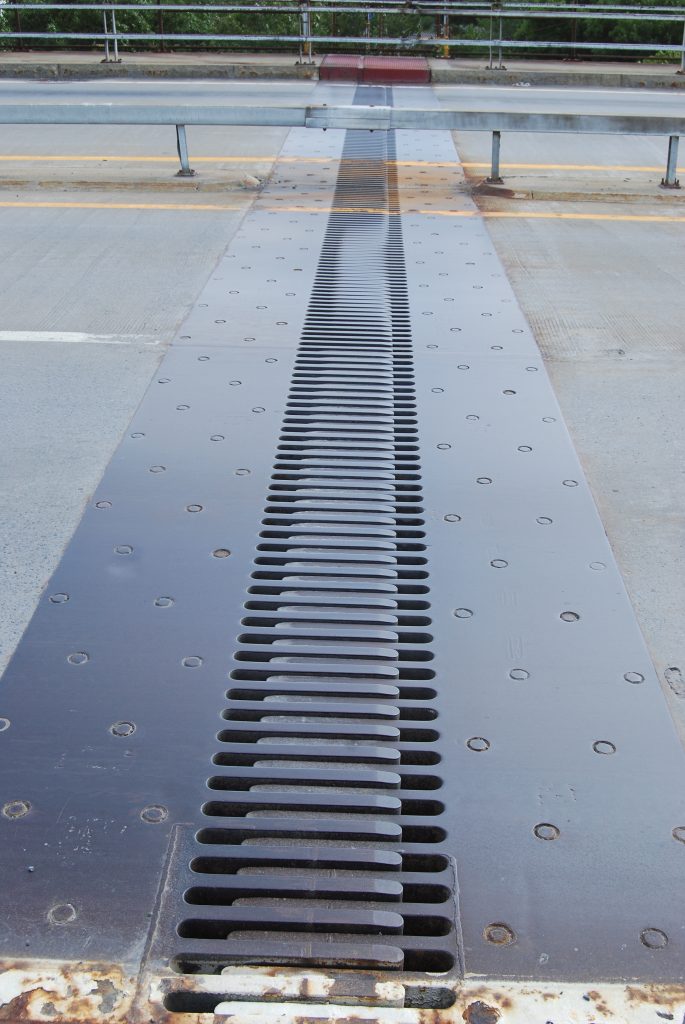 A photograph of a thermal expansion joint in a bridge. The expansion joint is a metal piece containing two sets of many interdigitated fingers with a gap in between.