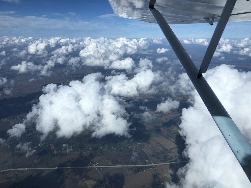 A photograph of cumulus clouds as seen out an airplane's window. The clouds separate a blue sky from farm fields in north central Illinois. The right wing and wing strut of the airplane can be seen out the window.