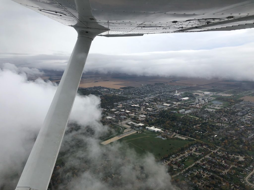 A photograph of stratus clouds as seen out an airplane's window. The clouds separate a gray sky from the city of DeKalb, Illinois. The right wing and wing strut of the airplane can be seen out the window.