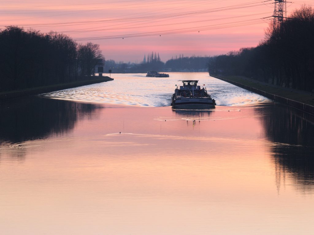 A barge traveling through a river generates bow waves. The river appears pink due to the setting sun. The riverbank is filled with trees. Powerlines can be seen crossing the river.