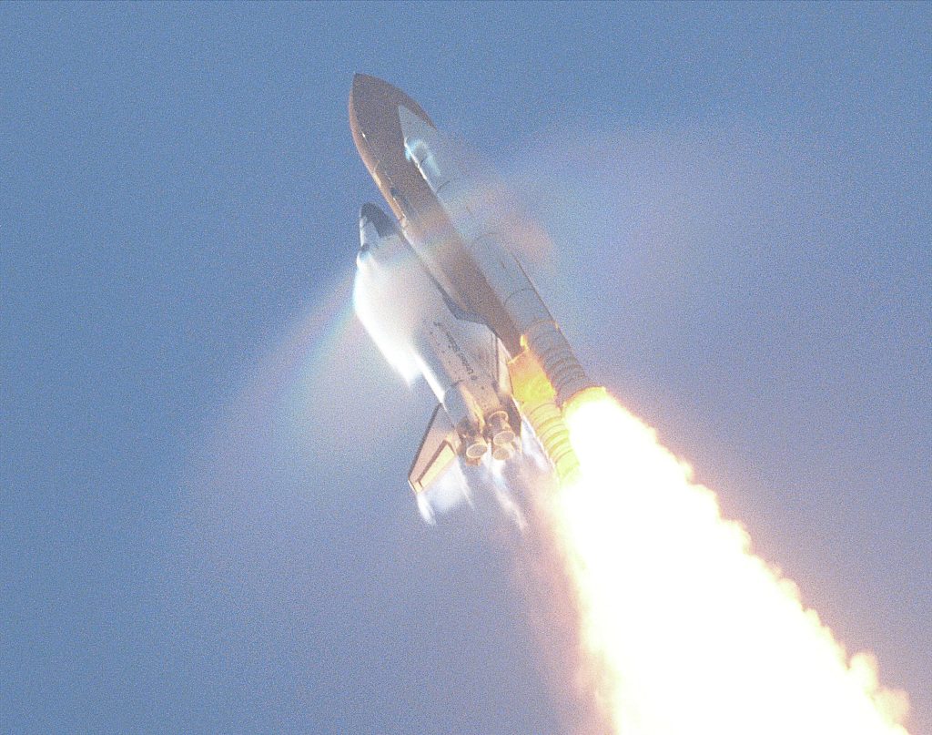The space shuttle taking off is viewed from below. Flames can be seen shooting out from the base of the solid rocket boosters. A cone shaped cloud appears emanating from the nose of the space shuttle. This is a shockwave.