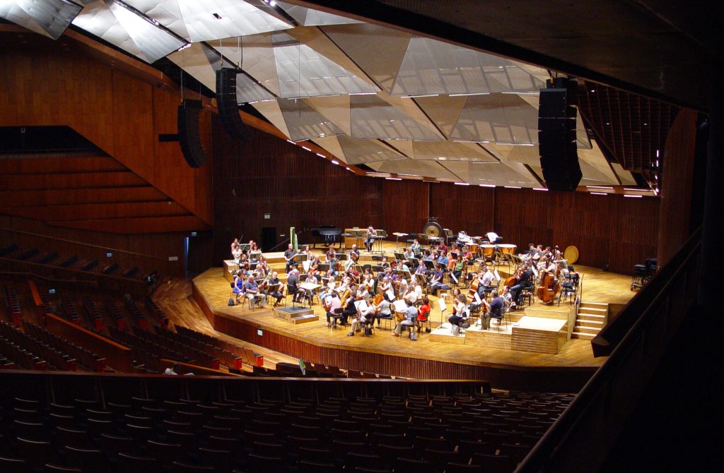 A photograph of an orchestra rehearsing in a concert hall. The entire hall can be seen, including the seats, stage, and ceiling. On the ceiling are many acoustic tiles positioned at different angles.