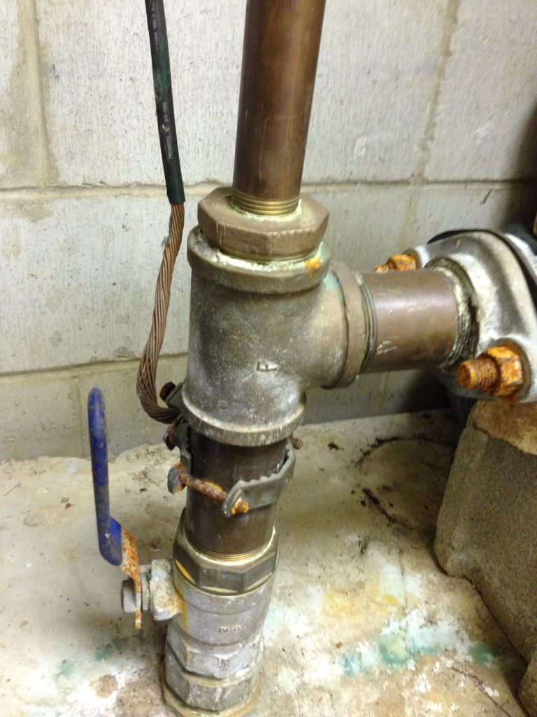 A photograph of a copper pipe plumbing system. There are a few pipes connected together that continue through the concrete floor, presumably into the Earth beneath. An electrical wire is connected to the plumbing, creating an Earth ground connection.