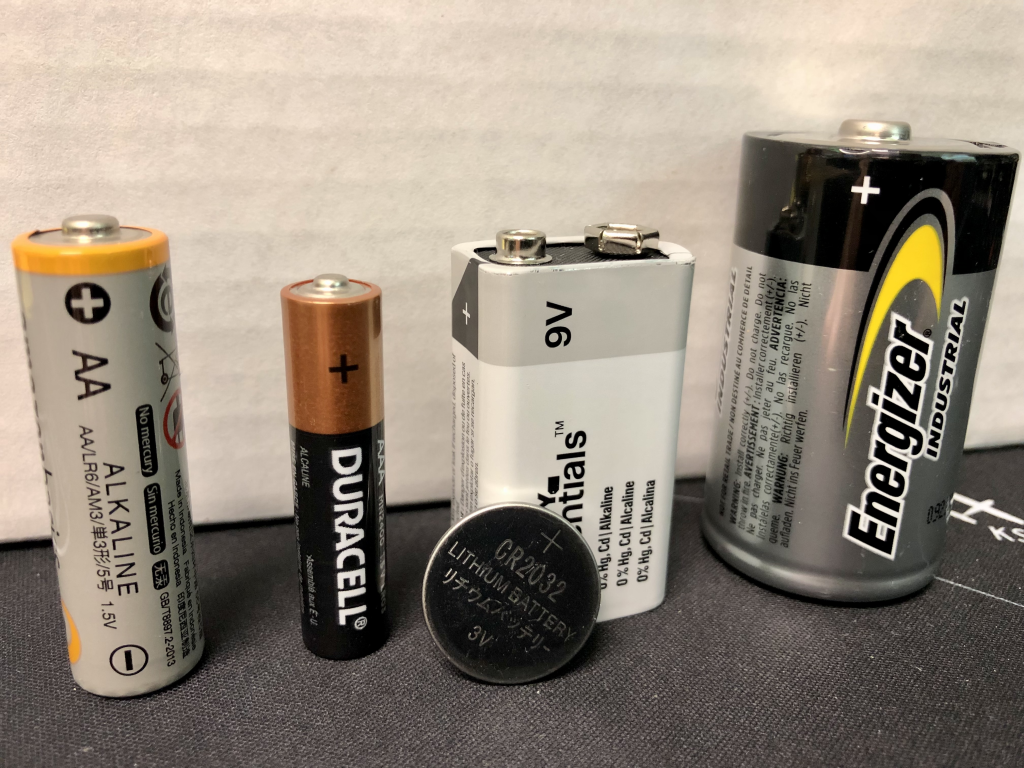 A photograph of several batteries. From left to right: AA, AAA, lithium coin cell, 9V, and D-cell battery.