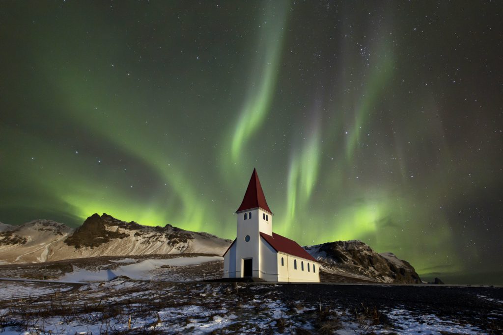 A photograph of the aurora borealis. They are wavy green and blue whisps seen in the atmosphere, surrounded by a night sky with some stars. The foreground of the photograph contains a white church on a snowy, rocky hill.