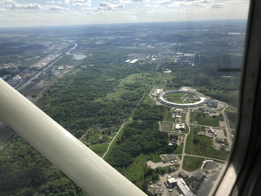 A photograph of the Advanced Photon Source as seen from out the window of an airplane. The building is a large toroidal-shaped white building.