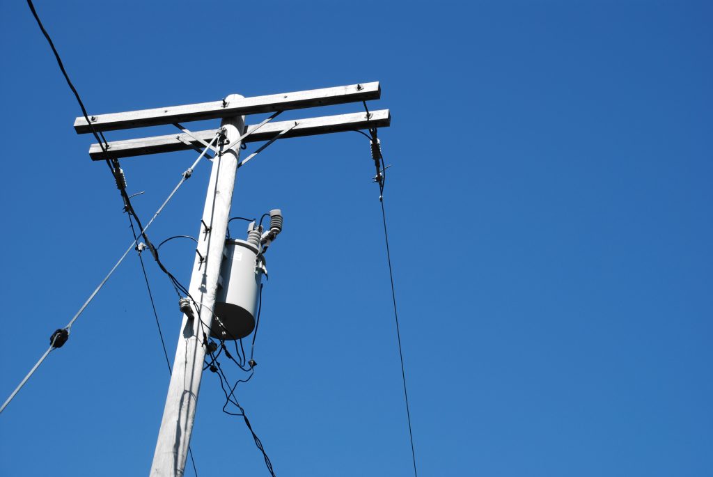 A photograph of a single distribution line tower, with a can-shaped transformer located partially down the pole. The photograph is shot from below and is silhouetted by a blue sky.