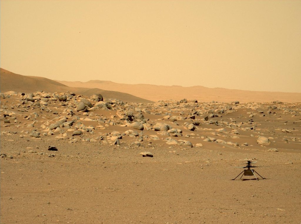 A photograph of Mars as taken from the Perseverance rover. The Ingenuity helicopter can be seen at the bottom right of the image, surrounded by the Martian terrain which appears to be sandy and rocky. The sky is a brownish pink color.