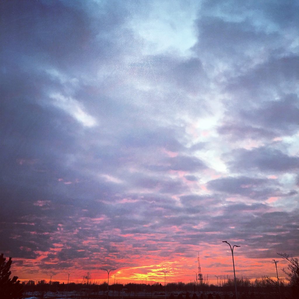 A photograph of sunset as taken from the College of DuPage parking lot. The sky is cloudy and appears with a gradient of colors, with bright red and orange toward the horizon, becoming blue father from the horizon. In the background some trees are silhouetted by the sky.