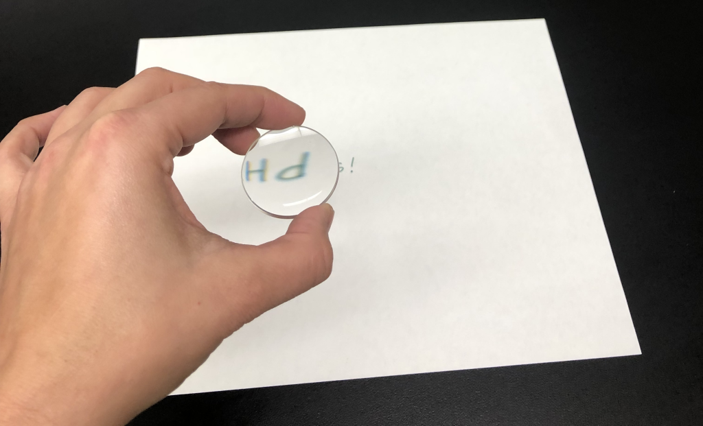 A photograph of Dr. Pasquale holding a lens in her hand. Through the lens can be seen the word "PHYSICS!", which is upside-down. In the background is the piece of paper with this word written on it. The object (the piece of paper with writing on it) is smaller than the image.
