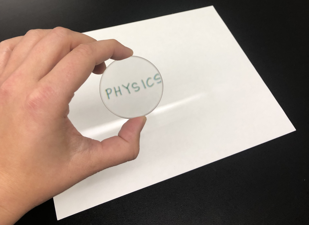 A photograph of Dr. Pasquale holding a lens in her hand. Through the lens can be seen the word "PHYSICS!" In the background is the piece of paper with this word written on it. The object (the piece of paper with writing on it) is smaller than the image.