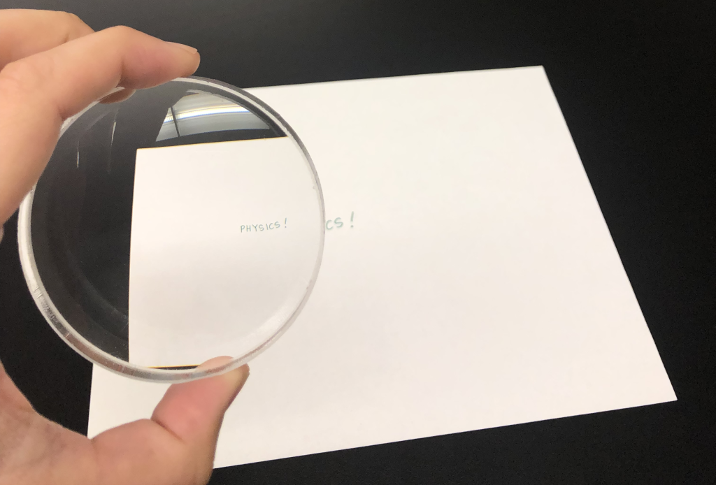 A photograph of Dr. Pasquale holding a lens in her hand. Through the lens can be seen the word "PHYSICS!" In the background is the piece of paper with this word written on it. The object (the piece of paper with writing on it) is larger than the image.