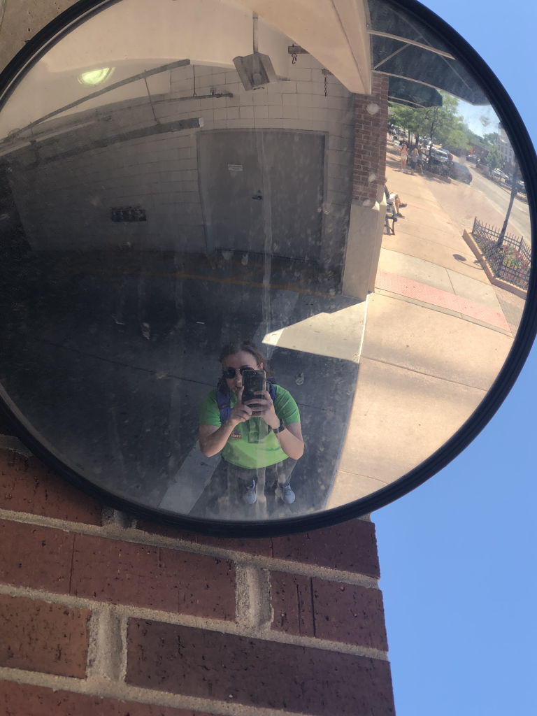 A photograph of a convex mirror connected to a brick wall on the corner of a parking garage. The reflection shows Dr. Pasquale, who appears much smaller than she is, as well as the surrounding sidewalk, adjacent roadway, and interior of part of the parking garage.