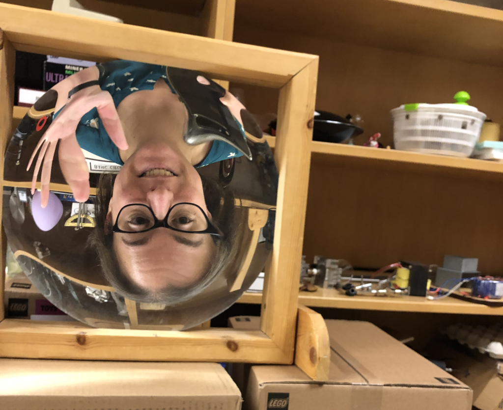 A photograph of Dr. Pasquale holding up her cellphone, taken in a concave mirror. She has short brown hair, is wearing eyeglasses and a green shirt. Her image appears larger than she is, is upside-down, and is slighly curved with the curvature of the mirror.