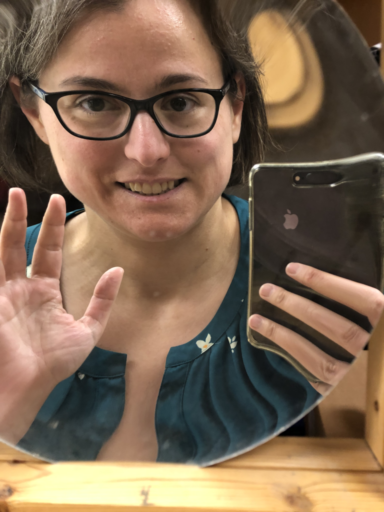 A photograph of Dr. Pasquale holding up her cellphone, taken in a concave mirror. She has short brown hair, is wearing eyeglasses and a green shirt. Her image appears larger than she is, and is slighly curved with the curvature of the mirror.