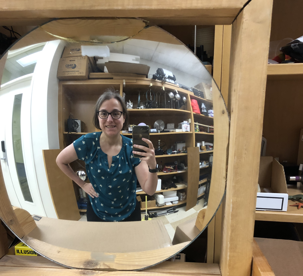 A photograph of Dr. Pasquale holding up her cellphone, taken in a convex mirror. She has short brown hair, is wearing eyeglasses and a green shirt. Her image appears smaller than she is, and is slighly curved with the curvature of the mirror.