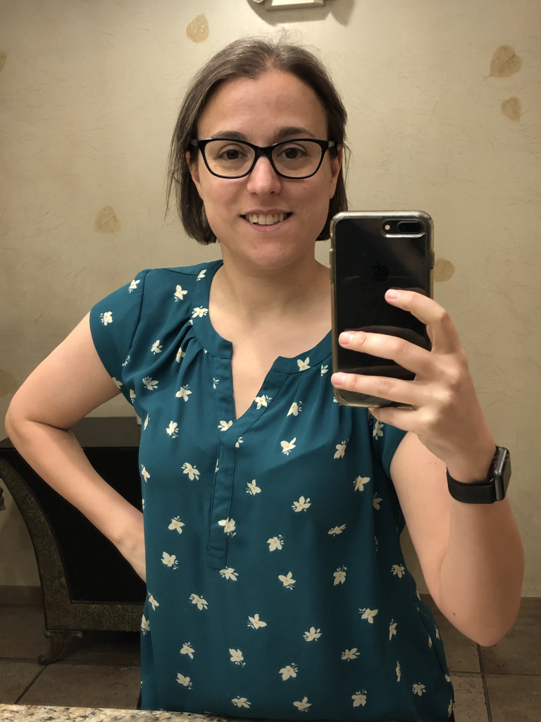 A photograph of Dr. Pasquale holding up her cellphone, taken in a planar mirror. She has short brown hair, is wearing eyeglasses and a green shirt.