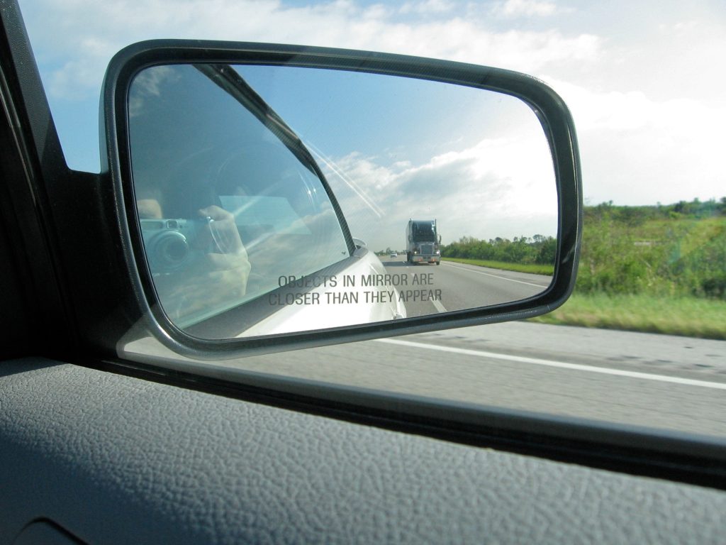 A photograph of a side mirror connected to a car. In the side mirror can be seen a truck traveling behind the car, as well as the highway that the car is driving on.