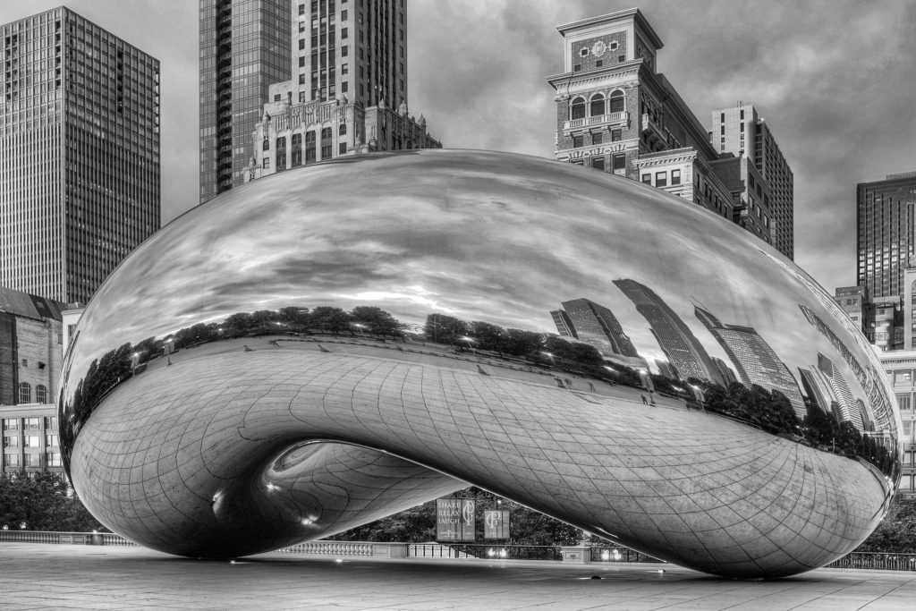 A photograph of the Cloud Gate sculpture in Chicago, Illinois. It is a giant, polished, smooth reflecting mirror shaped like a bean. In the background are buildings in the city. These can be seen reflected in the sculpture. Following the law of reflection, each reflection is upright and smaller than the object that created the reflection.