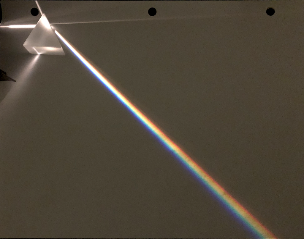 A photograph of a prism. White light enters from the left and is split as it exists the prism to the lower right at an angle. Red light bends the least and violet light bends the most.