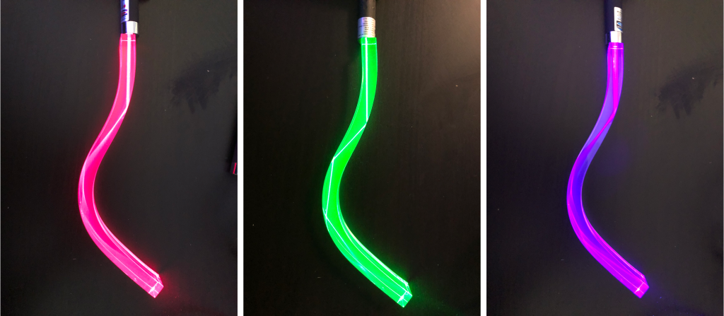 Three photographs. On the left is a photograph of a red laser pointer shining into a piece of curved plastic. Whenever the plastic curves, the light reflects at the plastic/air interface and remains internal to the plastic. In the middle is the same phenomenon with a green laser pointer. On the right is the same phenomenon with a blue laser pointer.