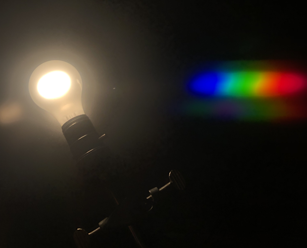 A photograph of an incandescent light bulb taken through a diffraction grating. The light bulb appears as a normal bulb that is on and shining white light. To the right is the area of constructive interference from the diffraction grating. Seen is a rainbow, with violet light closest to the bulb and red light farthest from the bulb.