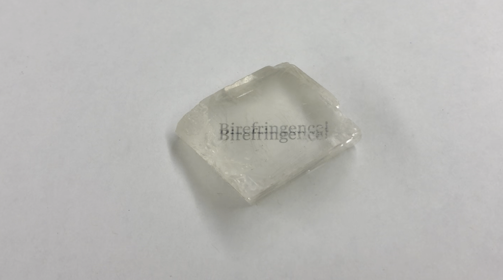 A photograph of a piece of quartz placed over the word "Birefringence!" written on a white piece of paper. The words appear as a double image, slightly separated from each other.