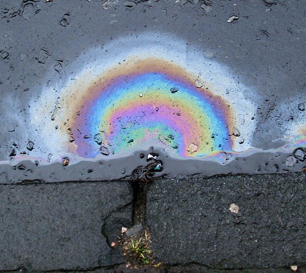 A photograph of a rainbow pattern caused in an oil slick resting on a puddle of water. In the background is asphalt