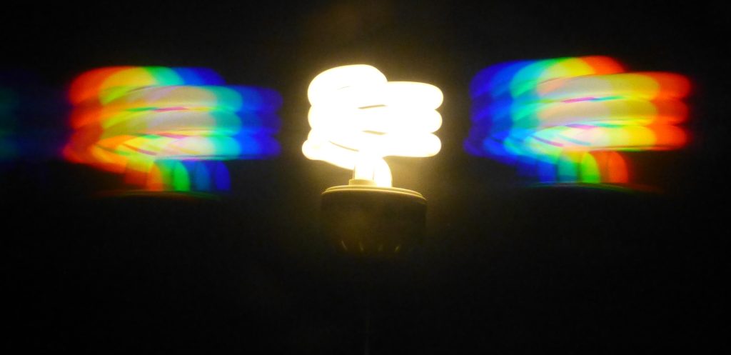 A photograph of a fluorescent bulb viewed through a diffraction grating. The bulb itself is glowing white. To each side (left and right) of the bulb can be seen the spectral components of light produced by the diffraction grating.